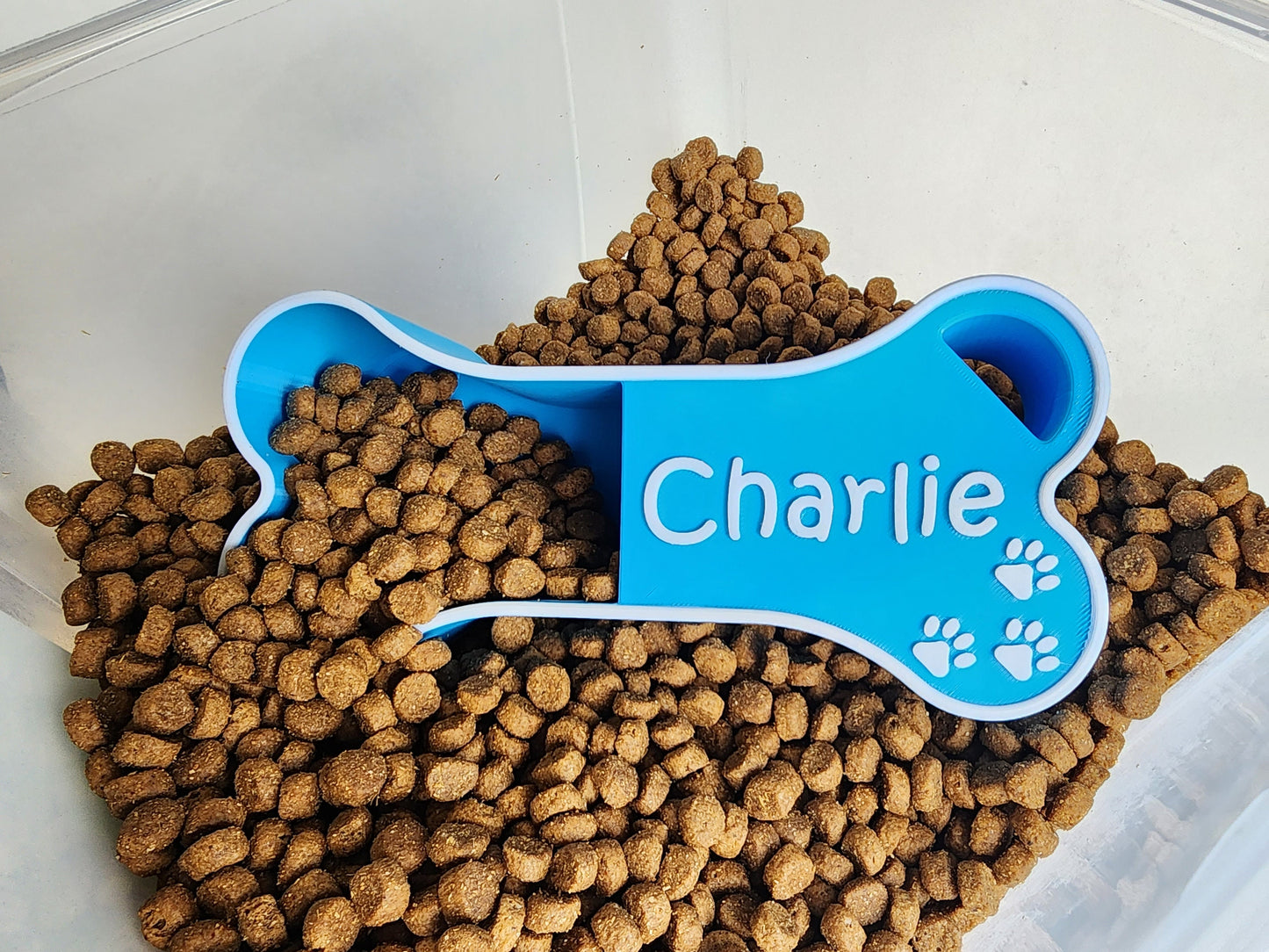 A light blue dog food scoop in the shape of a bone with the name Charlie in white. The scoop is in a container of dog food