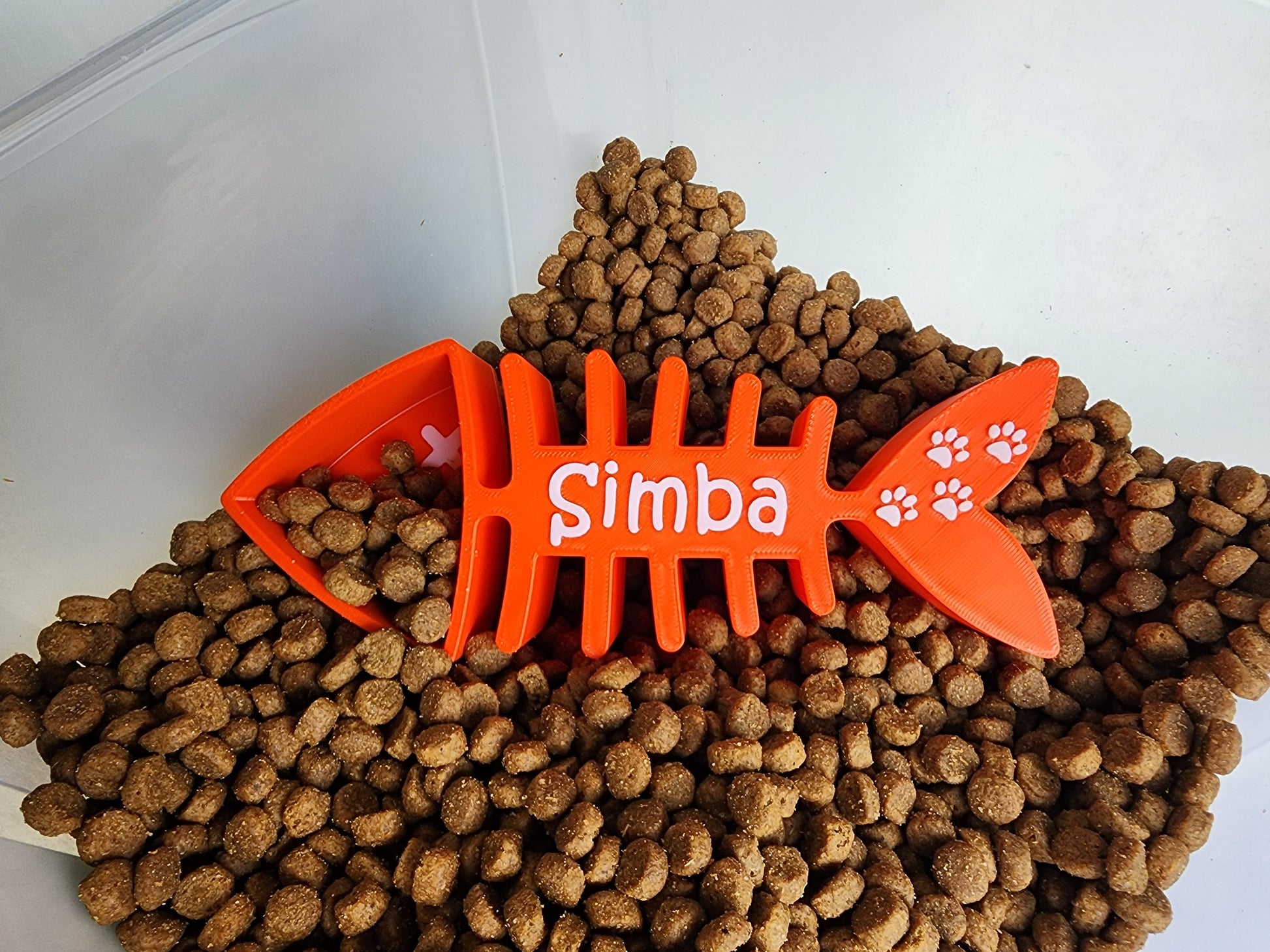 An orange 1/4 cup cat food scoop shaped like a fish skeleton with the name Simba and small paw prints in white. The scoop is in a container of cat food.