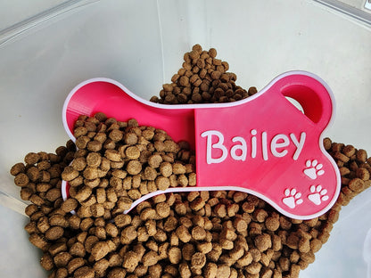 A pink dog food scoop in the shape of a bone with the name Bailey in white. The scoop is in a container of dog food