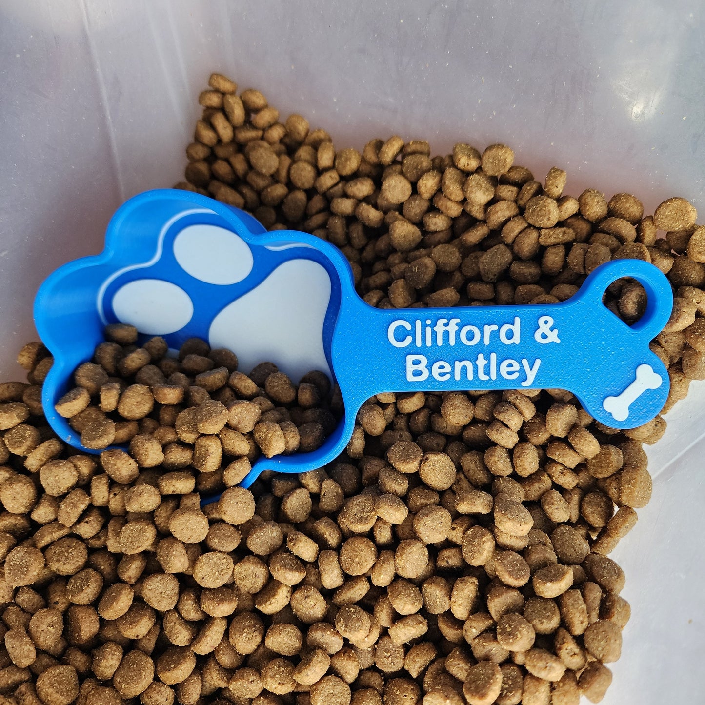 A blue 1 cup dog food scoop in the shape of a paw print with the names Clifford and Bentley in white. The scoop is in a container of dog food